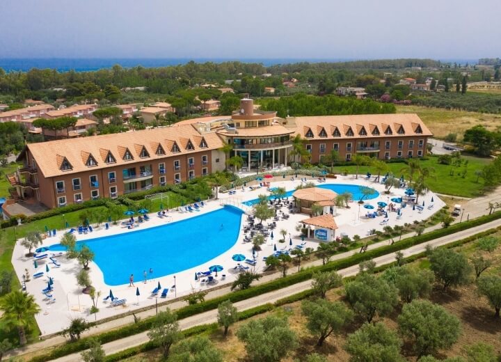 Immagine di un hotel con piscina, architettura calda e naturale, circondato da alberi e un paesaggio sereno.