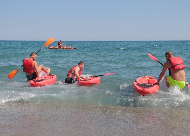 Quattro uomini in kayak su una spiaggia serena, indossando giubbotti di salvataggio rossi e remando in acque tranquille.