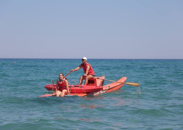 Due persone in una barca rossa nel mare blu, una seduta e l'altra in piedi con un remo, atmosfera serena.