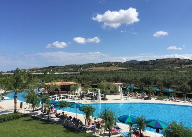 Una piscina blu circondata da verde lussureggiante, sedie a sdraio e colline di ulivi sotto un cielo sereno.