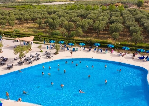 Una piscina blu circondata da sdraio e ombrelloni, immersa in un paesaggio verde, con persone che si rilassano e nuotano.