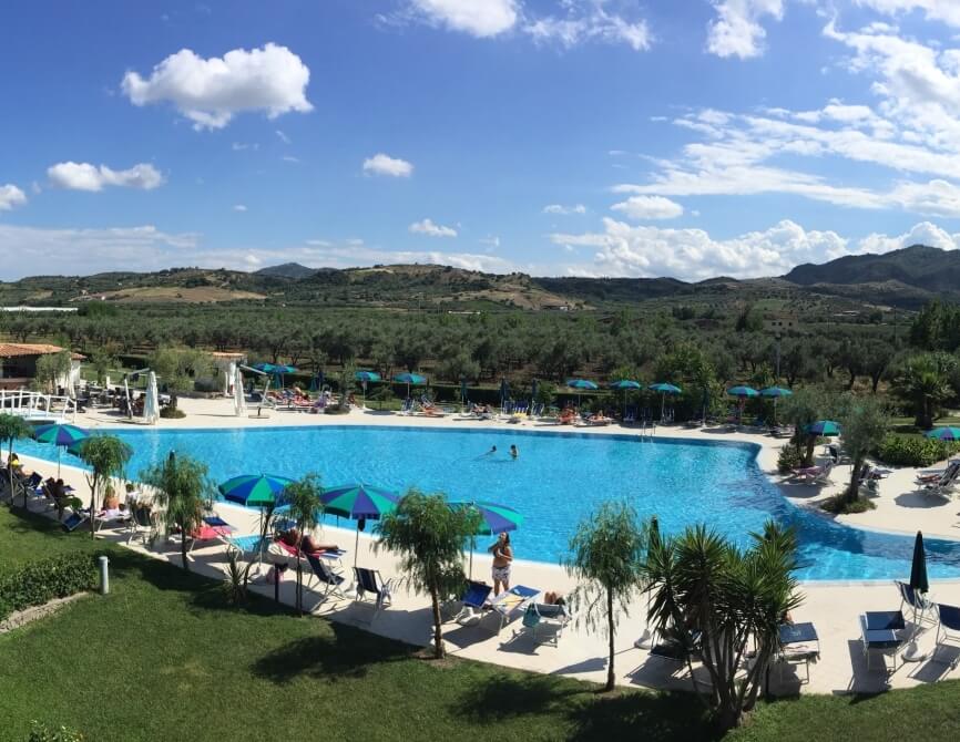 Una piscina all'aperto circondata da verde lussureggiante, con persone che si rilassano e nuotano sotto un cielo sereno.