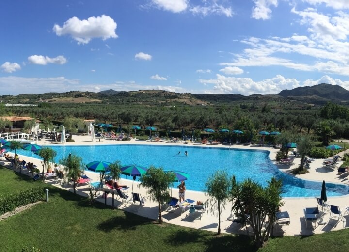 Una piscina serena con persone che si rilassano, circondata da un prato verde, palme e colline sotto un cielo blu.