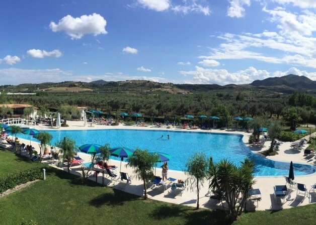 Una piscina serena circondata da palme, lettini e montagne, sotto un cielo blu con nuvole leggere.