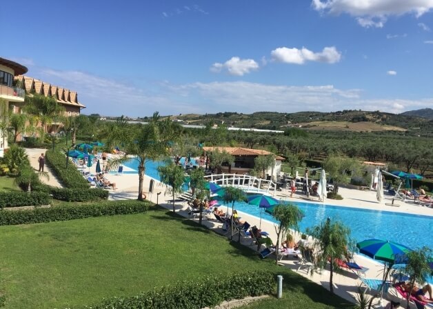 Una scena serena di un'area piscina con lettini, ombrelloni, un hotel e colline verdi sotto un cielo blu.