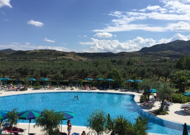 Una piscina ovale circondata da verde lussureggiante e montagne, con persone che si rilassano e nuotano sotto un cielo blu.