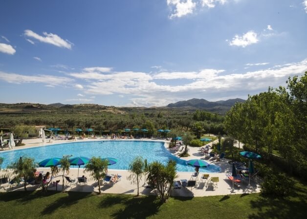 Una piscina all'aperto circondata da verde lussureggiante, montagne, ombrelloni colorati e un cielo blu sereno.