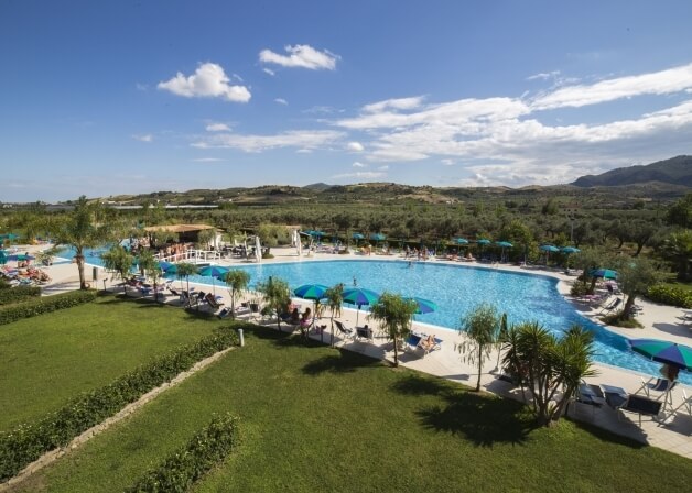 Una piscina serena circondata da sdraio, ombrelloni, piante e montagne, sotto un cielo blu con alcune nuvole.