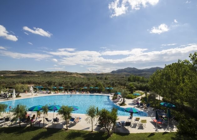 Una grande piscina rettangolare con sedie a sdraio e ombrelloni, circondata da alberi e colline sotto un cielo blu.