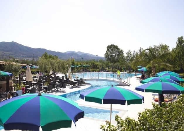 Impostazione serena a bordo piscina con ombrelloni, lettini, area pranzo e vista su alberi e montagne.