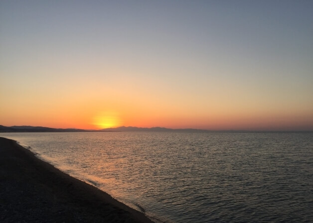 Tramonto su un corpo d'acqua calmo, con cielo blu e arancione, montagne in lontananza e una spiaggia sabbiosa.