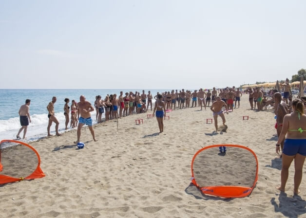 Una scena di spiaggia con persone che giocano e si godono il sole, attrezzature sulla sabbia e un'acqua calma sullo sfondo.