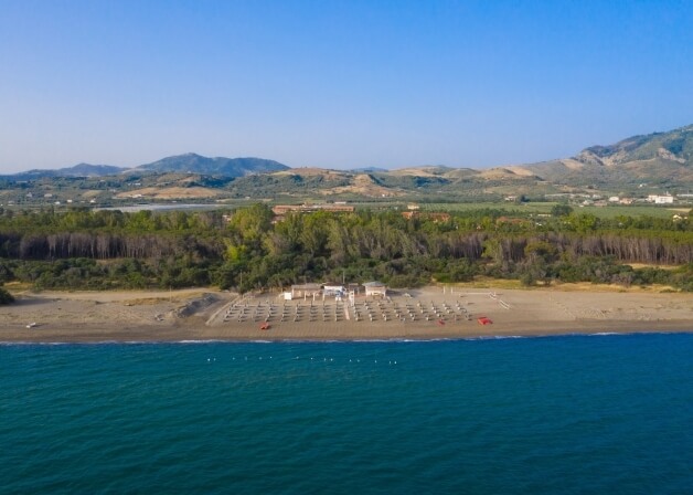 Una scena serena di spiaggia con acqua calma, sabbia, strutture bianche, oggetti rossi e colline verdi all'orizzonte.