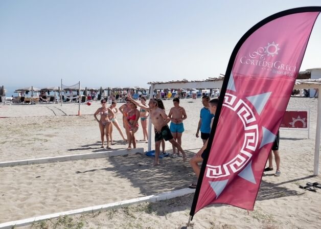 Spiaggia con una grande bandiera rosa "Corte del Greco" e persone in costume vicino a una rete da pallavolo.