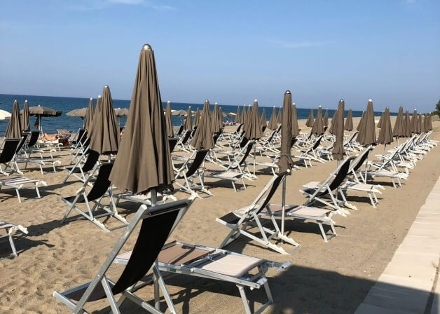 Spiaggia serena con lettini e ombrelloni vuoti, acque blu calme e cielo chiaro, senza presenza umana.