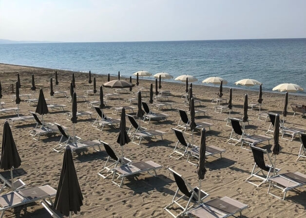 Spiaggia serena con lettini bianchi e marroni, ombrelloni chiusi, tavoli da picnic e onde dolci sotto un cielo blu.