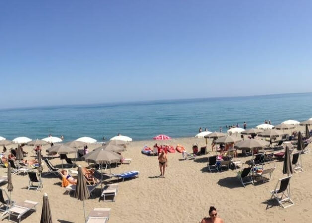 Una scena serena di spiaggia con sabbia, oceano calmo e bagnanti che si rilassano e giocano sotto un cielo blu.