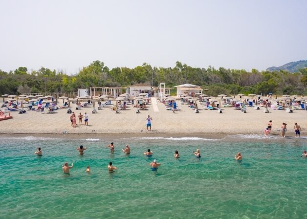 Scena di spiaggia con persone che nuotano e prendono il sole, ombrelloni, sabbia, foresta e montagne sullo sfondo.