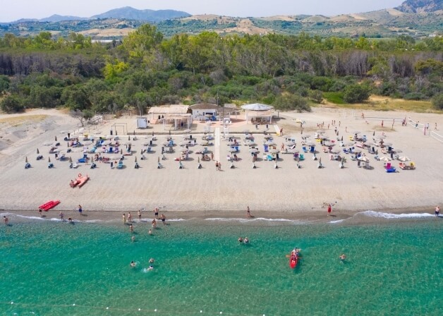 Spiaggia affollata con persone che nuotano e prendono il sole, ristorante sullo sfondo e montagne in lontananza.