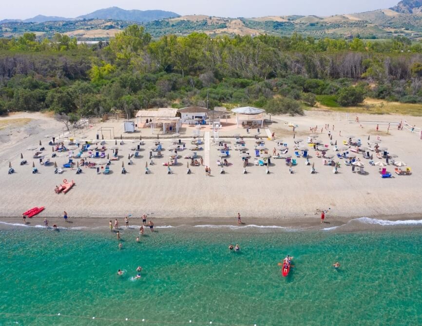 Vista aerea di una spiaggia affollata con ombrelloni, persone in acqua e colline sullo sfondo. Atmosfera di relax e divertimento.