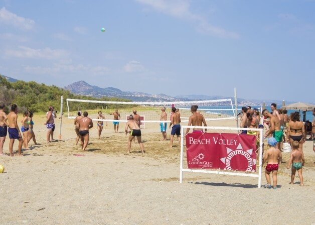 Giocatori di beach volley in azione su una spiaggia, con alberi, montagne e acqua calma sullo sfondo.