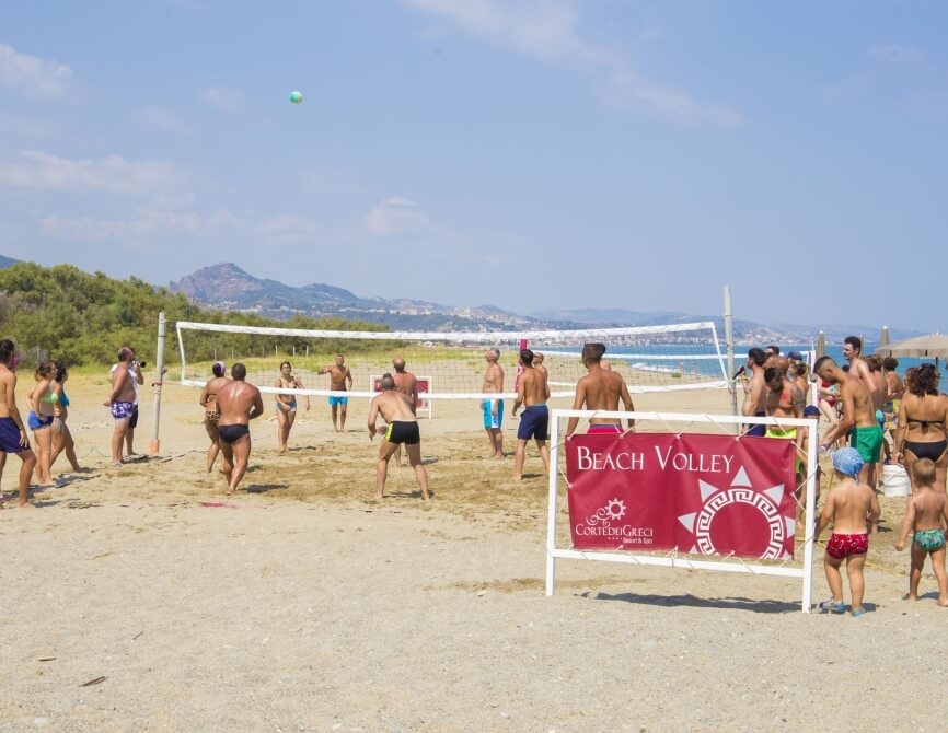 Giocatori di pallavolo sulla spiaggia con un banner "BEACH VOLLEY" e un bar sullo sfondo di un paesaggio sereno.