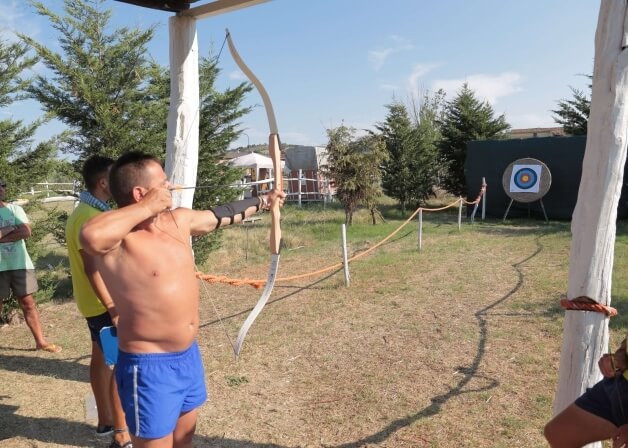 Un arciere in pantaloni blu mira a un bersaglio, osservato da due persone, in un campo di tiro all'aperto.