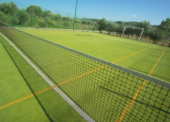 Campo sportivo all'aperto con rete da tennis, sezione per tennis a sinistra e campo da calcio a destra, sotto un cielo blu.