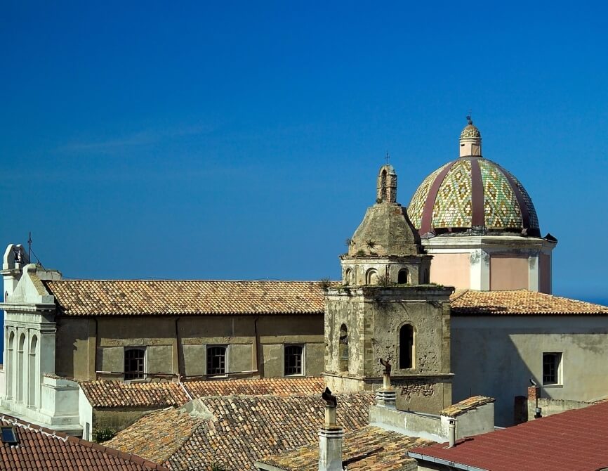 Panorama di una città con una cattedrale centrale, cupole verdi, tetti in terracotta e un murale sbiadito. Cielo blu.