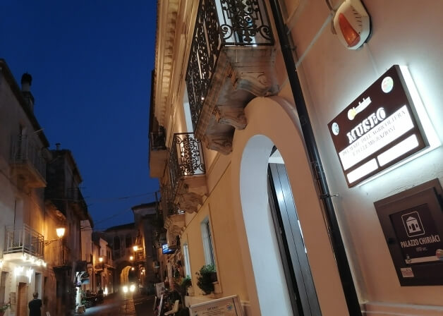 Strada notturna con segno "MUSEO", orologi e edifici storici con balconi e illuminazione.