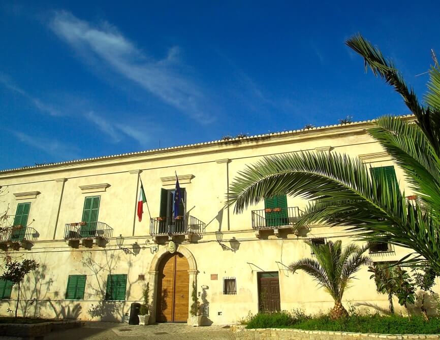 Edificio grande di colore beige con bandiera italiana, finestre con persiane verdi e sentiero circondato da piante e palme.