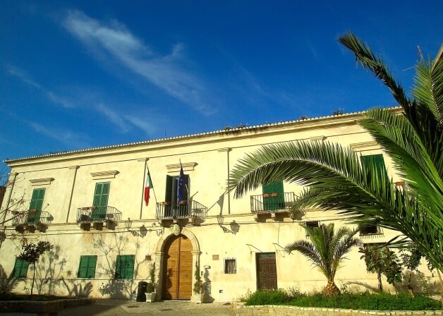 "Edificio in stile classico con porta ad arco, balcone e bandiere italiana e dell'UE, immerso in un giardino con palme e cielo a