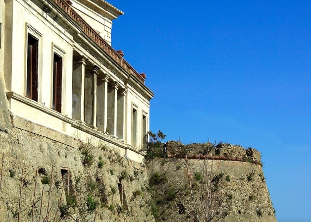 Edificio storico bianco con tetto in mattoni rossi, colonne e muro di pietra, sotto un cielo blu sereno.