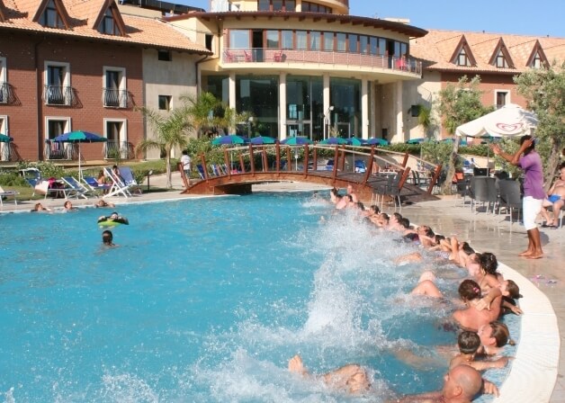 Una piscina all'aperto affollata con persone che si rilassano, un giovane che salta, e un hotel reddish-tan sullo sfondo.