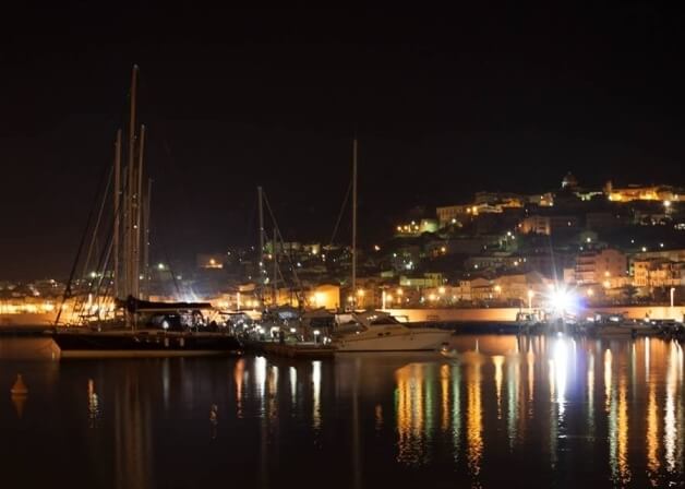 Una scena notturna serena di un marina con barche a vela illuminate, riflettendosi nell'acqua calma e circondata da edifici.