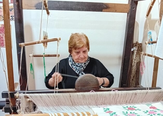 Donna al telaio in legno, indossa un top nero e una sciarpa blu e bianca, mentre tesse un tessuto bianco con fiori rosa.