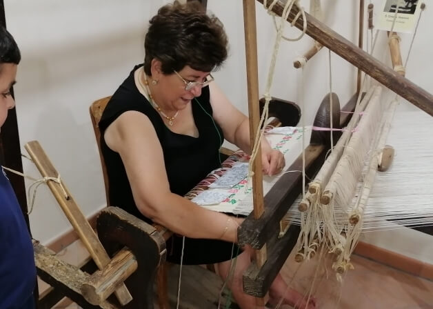 Donna che tesse su un telaio di legno, indossando un vestito nero, con un progetto di tessitura davanti a lei.