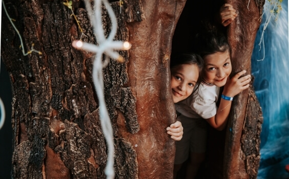Due bambini si affacciano dietro una grande struttura alberata illuminata.
