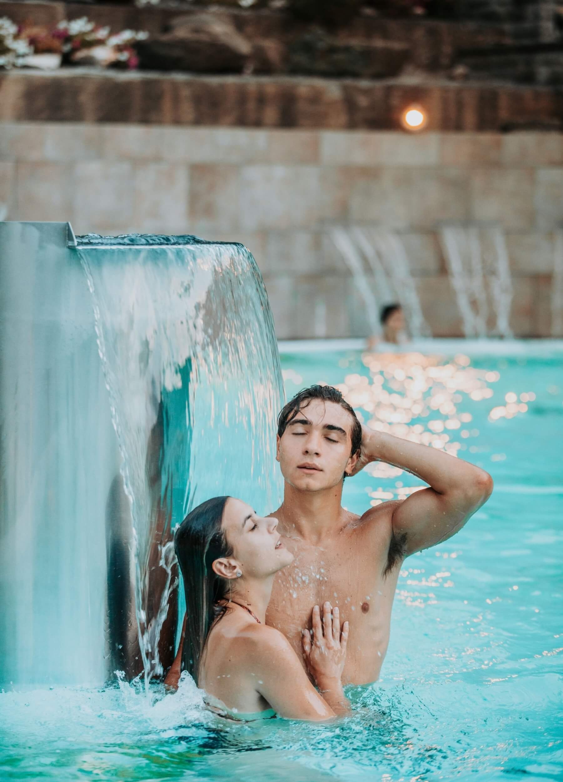 Uomo e donna in una piscina d'acqua turchese accanto a una cascata metallica.