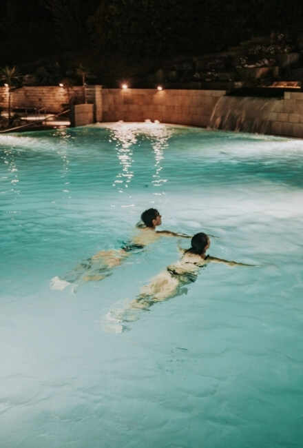 Due persone nuotano in una piscina turchese illuminata di notte.