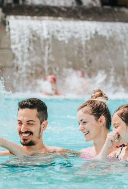 Uomo e due donne sorridono in piscina con cascata sullo sfondo.