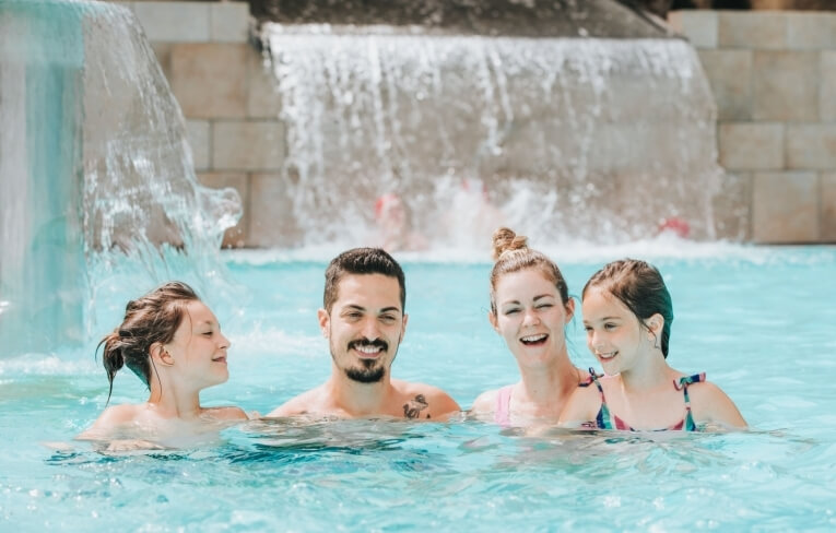 Una famiglia di quattro persone gioca in piscina con cascate sullo sfondo.