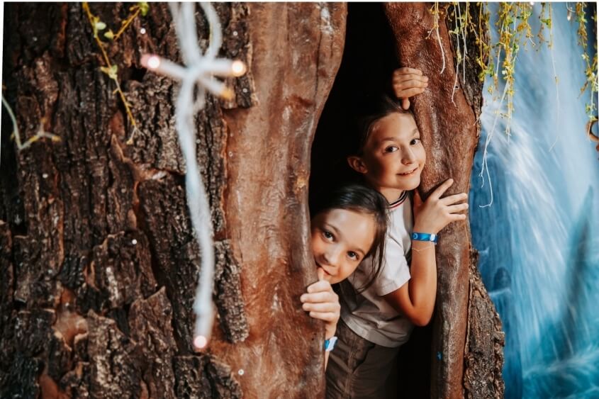 Due bambini guardano da un tronco d'albero decorato con luci.