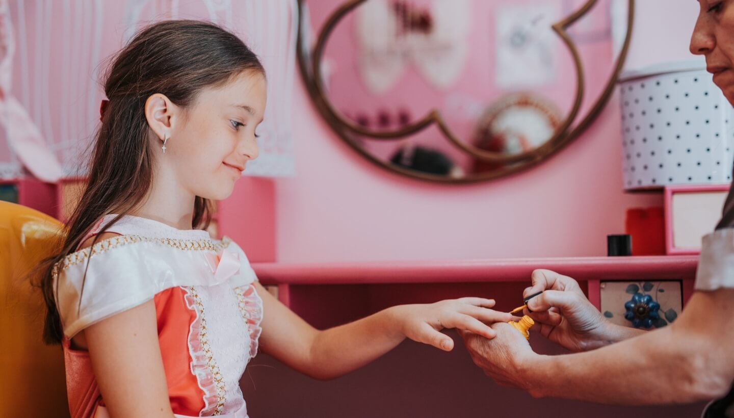 Bambina in abito rosa mentre le si applica smalto giallo.