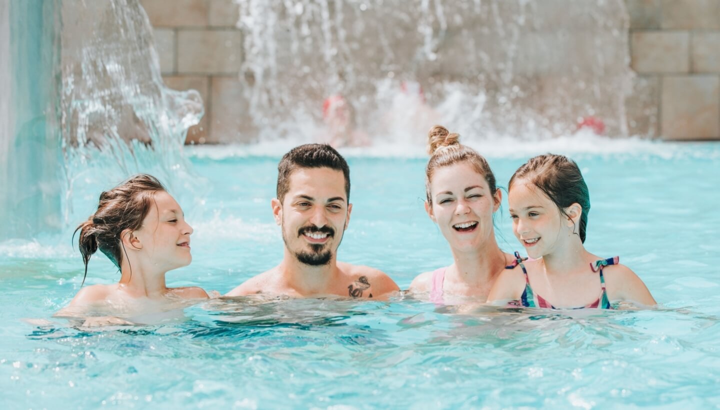 Una famiglia di quattro persone gioca in piscina con acqua che scorre sullo sfondo.