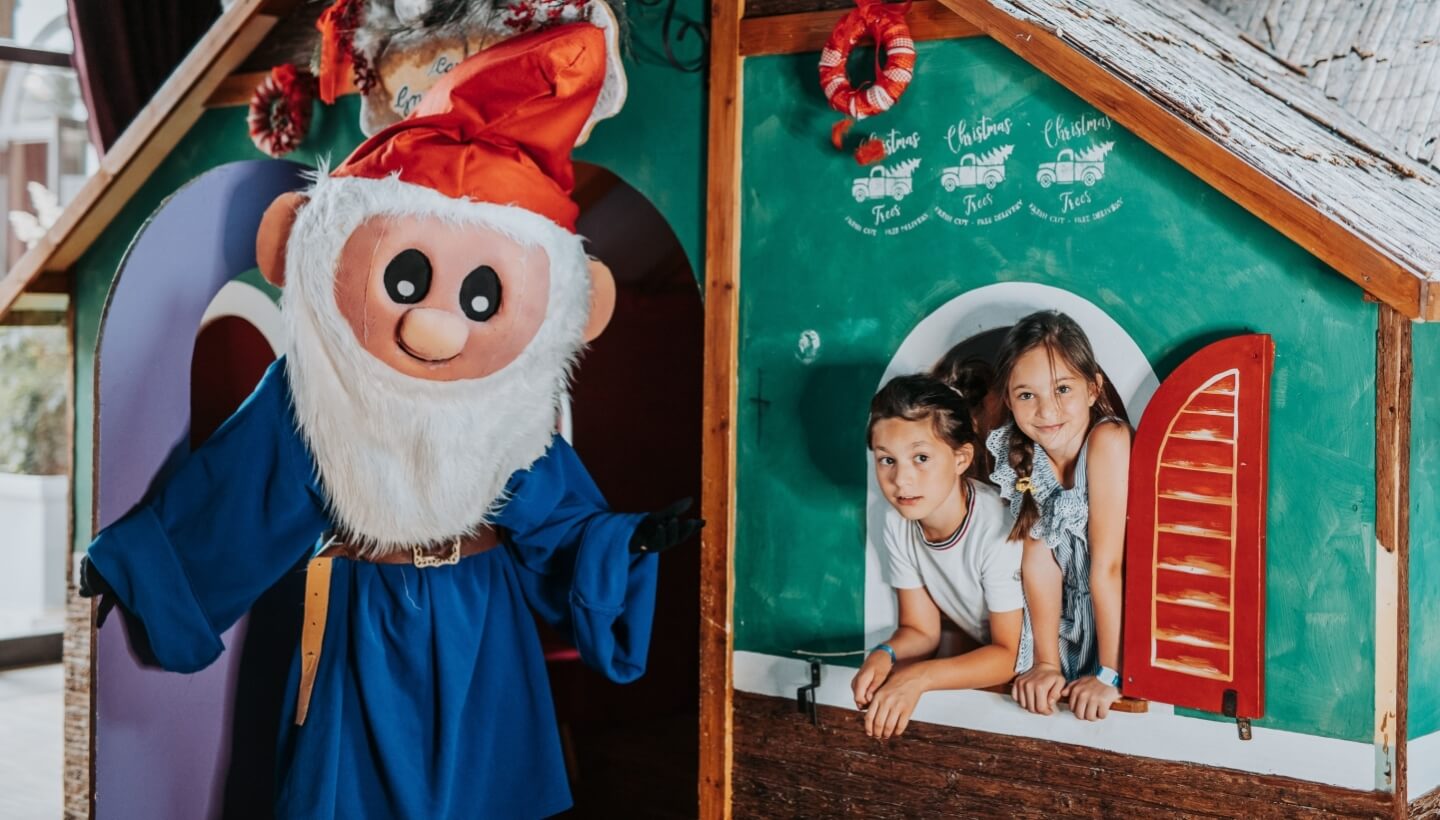 Costumed figure with children in playhouse, "Christmas Trees" sign visible.   Figura in costume con bambini nella casa, visibile il cartello "Alberi di Natale".
