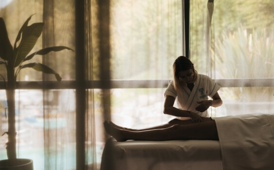 Person in white robe massages leg of another on a table.   Eine Person in einem weißen Gewand massiert das Bein einer anderen.