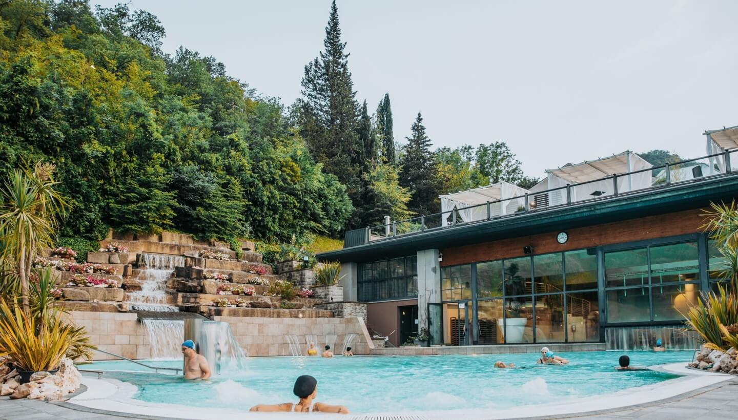 Piscina all'aperto con persone, cascata e edificio moderno sullo sfondo.