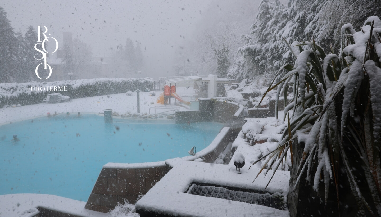 Piscina all'aperto innevata con persone, scivolo e piante.