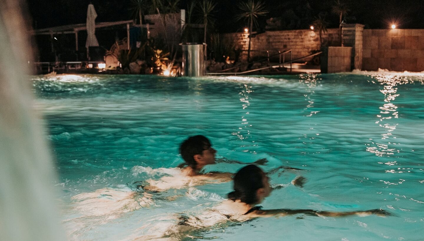Due persone nuotano in una piscina illuminata di notte.